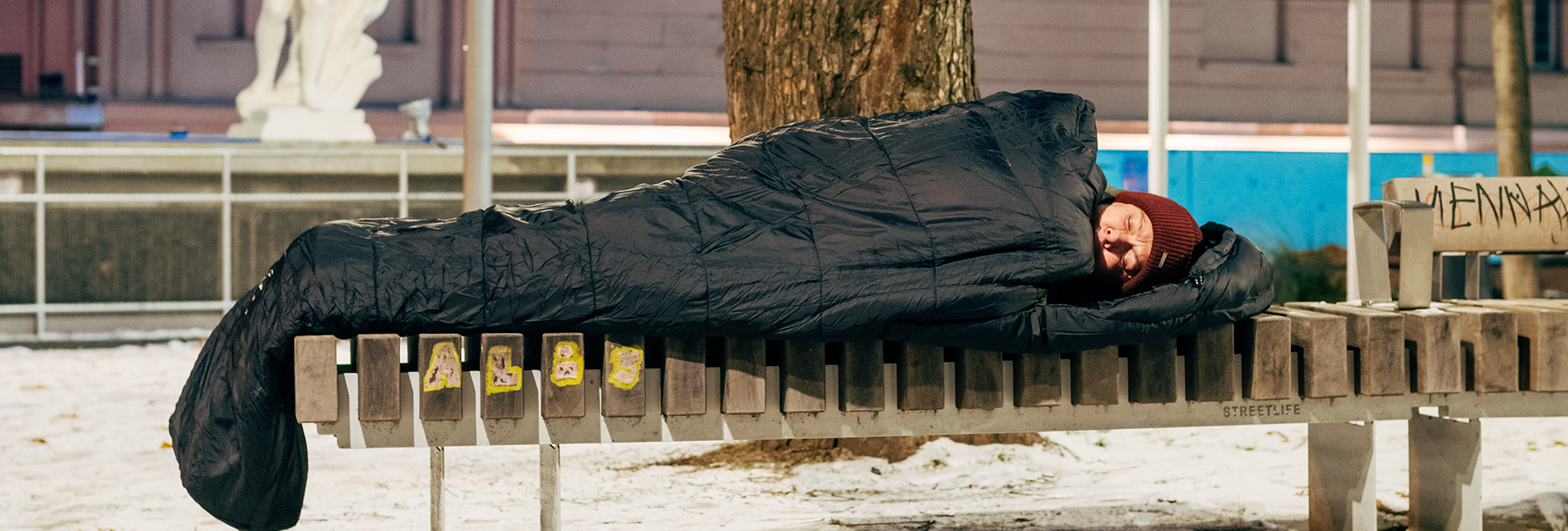 Wohnungslose Person schläft in einem Schlafsack auf einer Bank im Winter